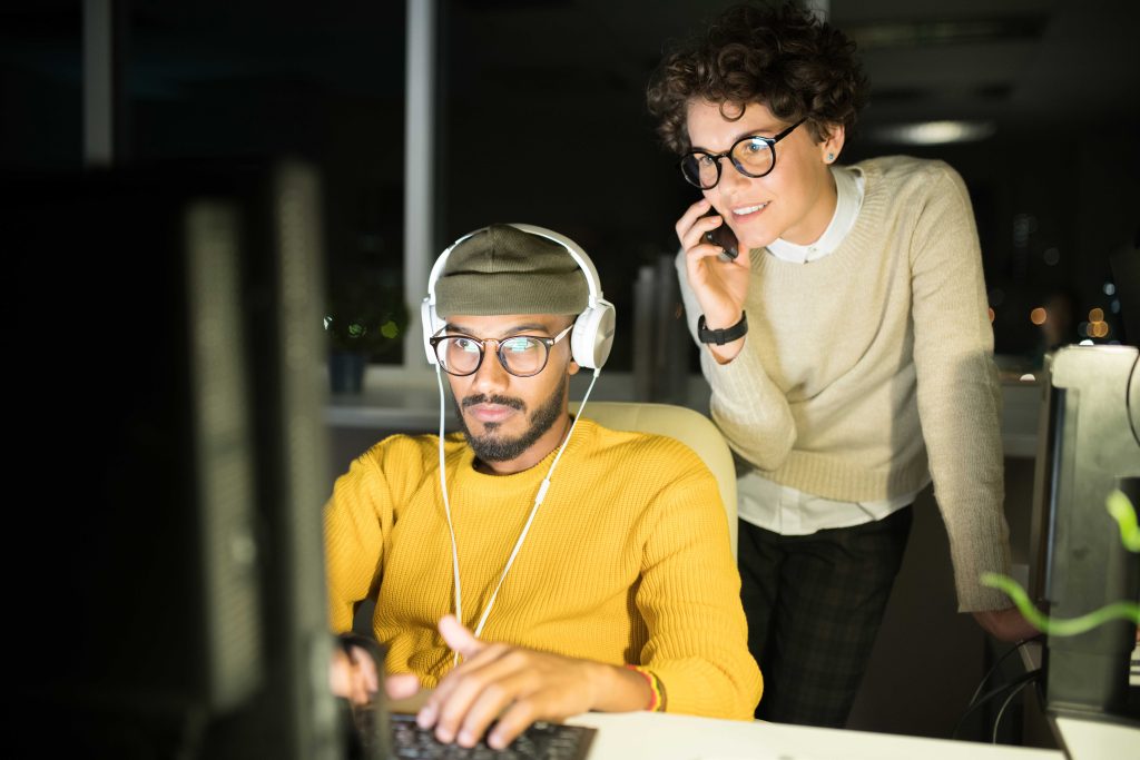 Portrait of Middle-Eastern computer programmer typing in dark office with manager watching him and speaking by phone